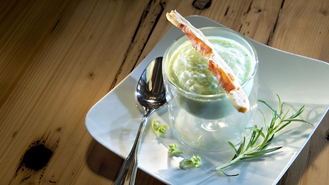 Ein grüner Dessertbecher mit einer cremigen Füllung steht auf einem weißen Teller. Daneben liegt ein Löffel und frische Kräuter sind dekorativ platziert. | © A.M.J. Hotel KG