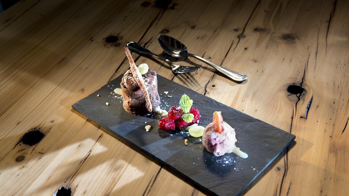 Ein elegantes Dessert auf einem schwarzen Servierbrett. Es besteht aus verschiedenen Eissorten, frischen Beeren und einer dekorativen Garnitur. | © A.M.J. Hotel KG