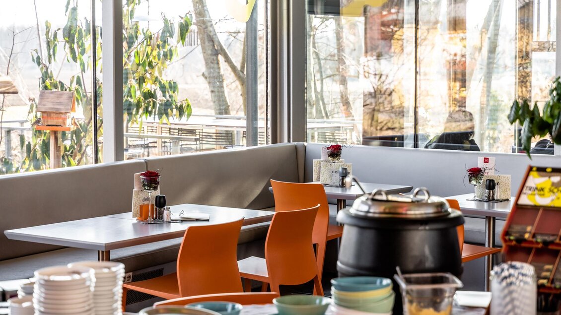 Ein modernes Restaurant mit großen Fenstern und gemütlichen Tischen. Es gibt orangefarbene Stühle und eine Auswahl an Geschirr auf den Tischen. | © Stephan Friesinger