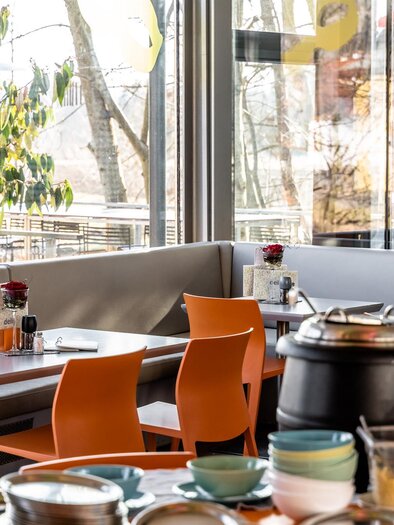 Ein modernes Restaurant mit großen Fenstern und gemütlichen Tischen. Es gibt orangefarbene Stühle und eine Auswahl an Geschirr auf den Tischen. | © Stephan Friesinger
