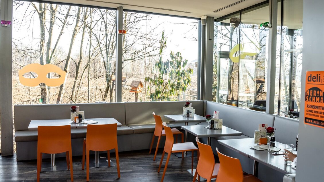 Ein helles Restaurant mit großen Fenstern und Blick auf die Natur. Die Tische sind modern eingerichtet und mit orangefarbenen Stühlen ausgestattet. | © Stephan Friesinger