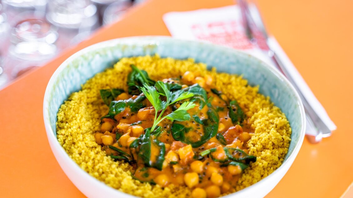 Eine Schüssel mit Couscous, Garbanzobohnen und Gemüse. Das Gericht ist mit frischen Kräutern garniert und auf einem orangefarbenen Tisch serviert. | © Stephan Friesinger