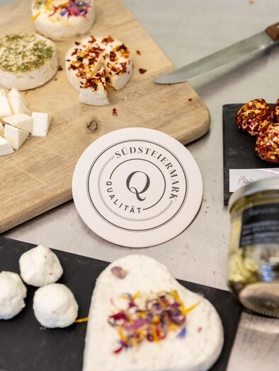 A selection of different types of cheese and small cheese balls on a wooden board and black plates. Additionally, there is a glass and a package of cheese on the table. | © @albrecherhof