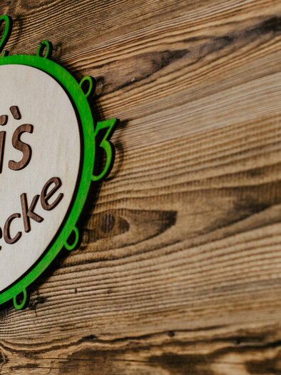 A wooden sign with the inscription "Dani's Genussecke". The sign features green and white colors and is mounted on a rustic wall. | © TV Region Graz - Mias Photoart