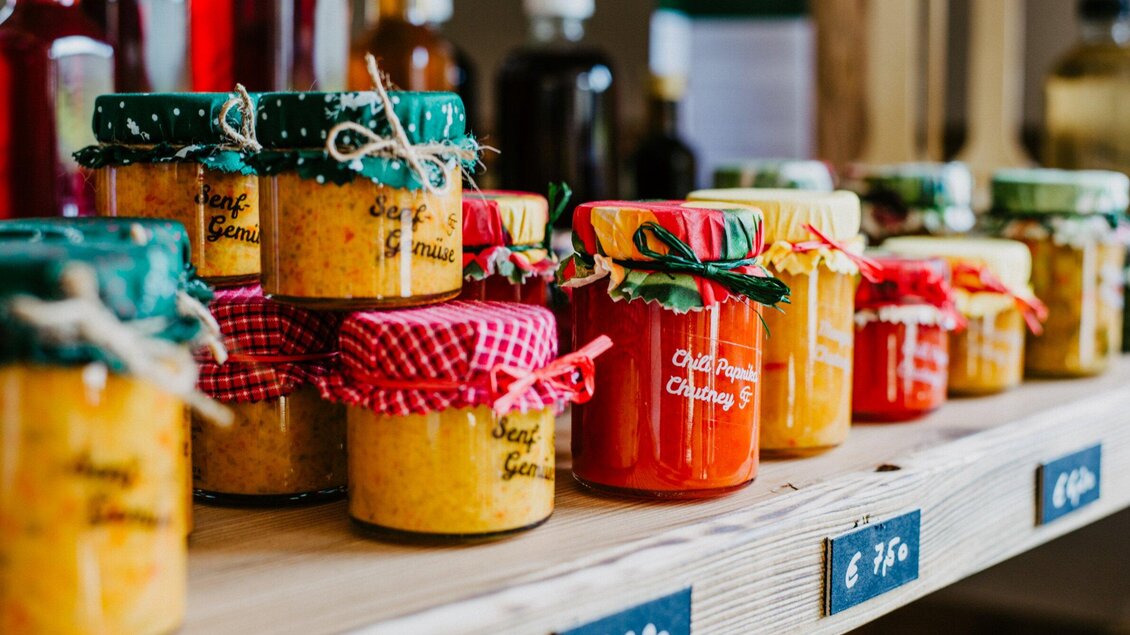 Bunte Gläser mit verschiedenen Marmeladen stehen auf einem Holztisch. Jedes Glas ist schön verpackt und dekoriert. | © TV Region Graz - Mias Photoart