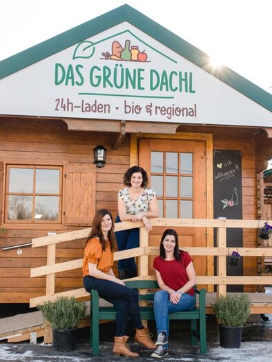 A wooden building with an inviting entrance and a sign that says "The Green Roof". Three women are sitting on a bench in front of the store, surrounded by plants. | © Das Grüne Dachl Gesbr