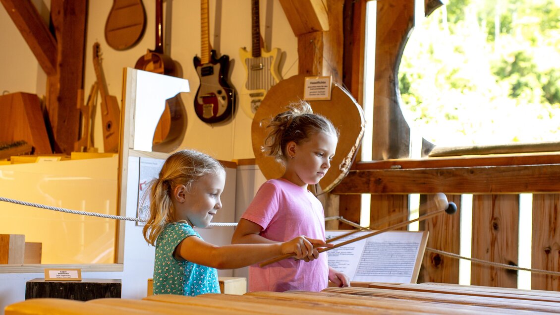Zwei Kinder spielen in einem Musikraum mit einem Schlaginstrument. Im Hintergrund sind verschiedene Musikinstrumente sichtbar.