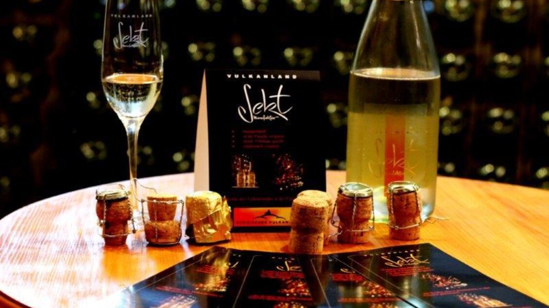 Ein elegantes Setup mit einem Glas Sekt, einer Flasche Sekt und zum Feiern vorbereiteten Korken. Umgeben von informativen Karten über die Sektauswahl. | © Vulkanlandsekt Manufaktur