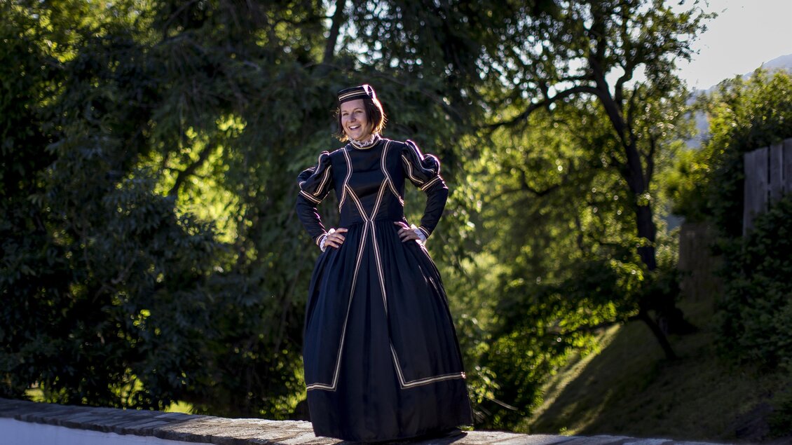 Eine Person in einem historischen schwarzen Kleid steht auf einer Mauer. Im Hintergrund sind grüne Bäume und eine sonnige Umgebung sichtbar. | © Holzwelt Murau