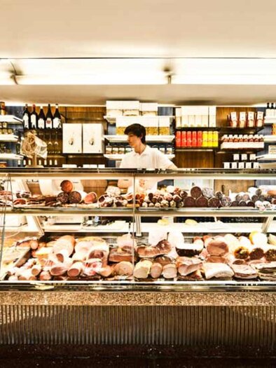 Große Glastheke voller Wurst- und Käsesorten, dahinter Mitarbeiter und Regale mit Wein und Konserven | © Frankowitsch