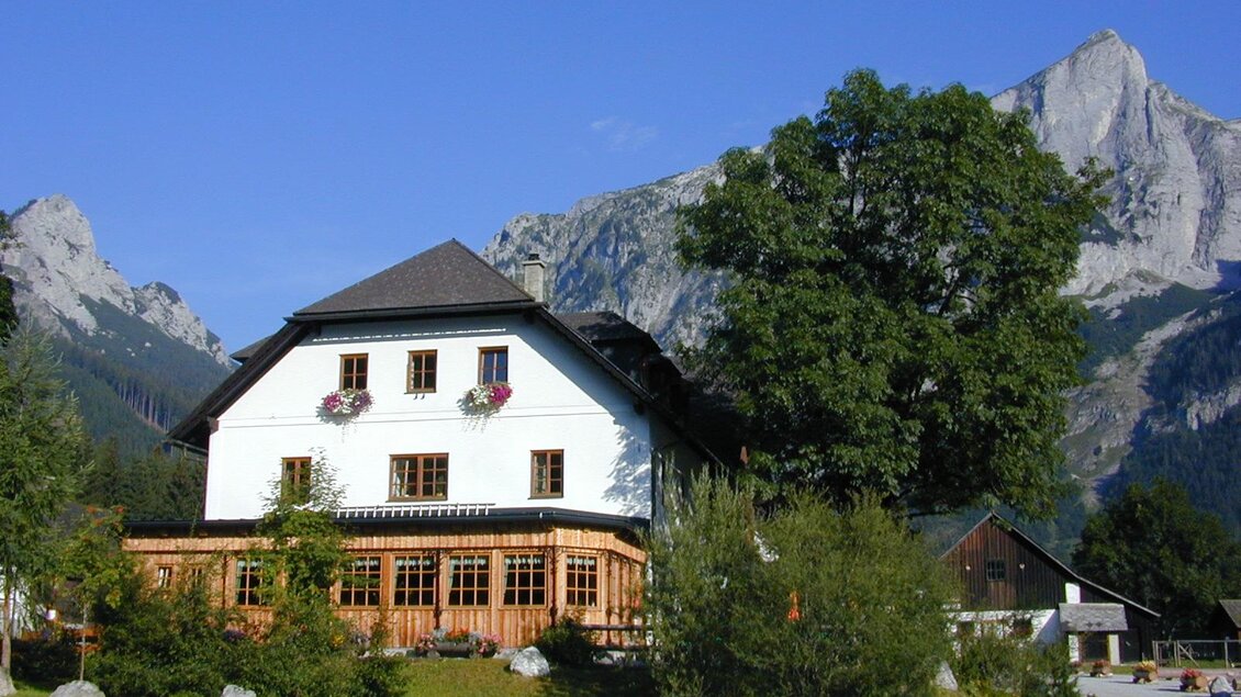 Ein malerisches Haus mit Holzveranda, umgeben von Bäumen und beeindruckenden Bergen. Der klare blaue Himmel ergänzt die idyllische Landschaft. | © ZWHS