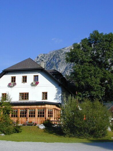 A picturesque house with a wooden porch, surrounded by trees and impressive mountains. The clear blue sky enhances the idyllic landscape. | © ZWHS