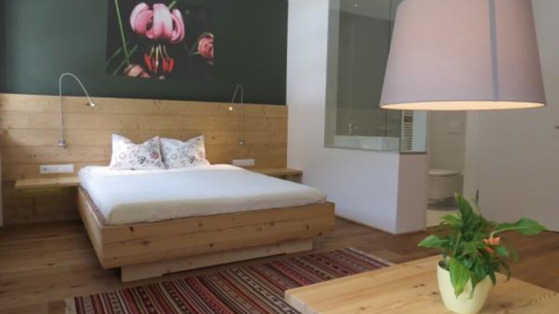 Ein modernes Schlafzimmer mit einem Holzbett und bunten Kissen. Die Wände sind in sanften Farben gehalten, und ein großer Lampenschirm sowie ein Pflanze auf dem Tisch sorgen für kuschelige Atmosphäre. | © ZWHS