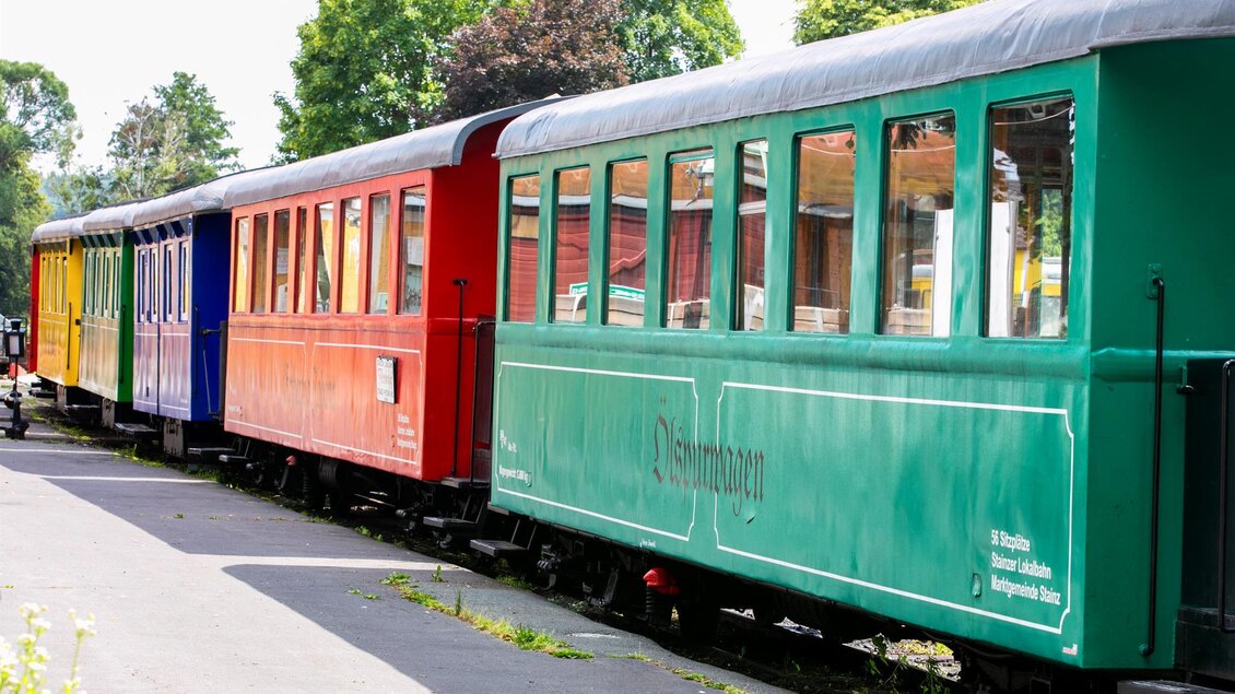 Eine bunte Reihe von Eisenbahnwagen steht an einem Bahnsteig. Die Wagen sind in verschiedenen Farben gestrichen und von Bäumen umgeben. | © Foto Augenblick