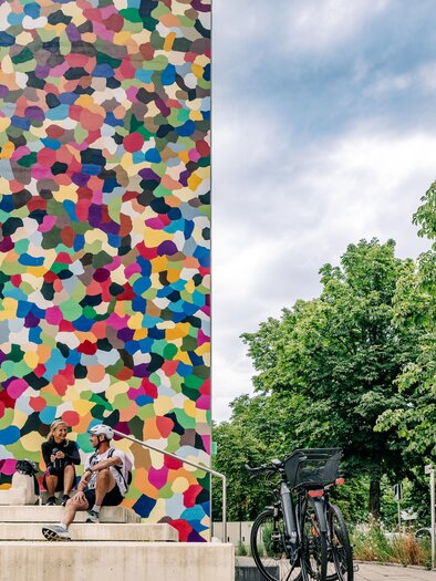 Two cyclists are taking a break in front of the most colorful church tower in the world in Feldbach in the Thermal & Volcano Land, Styria.