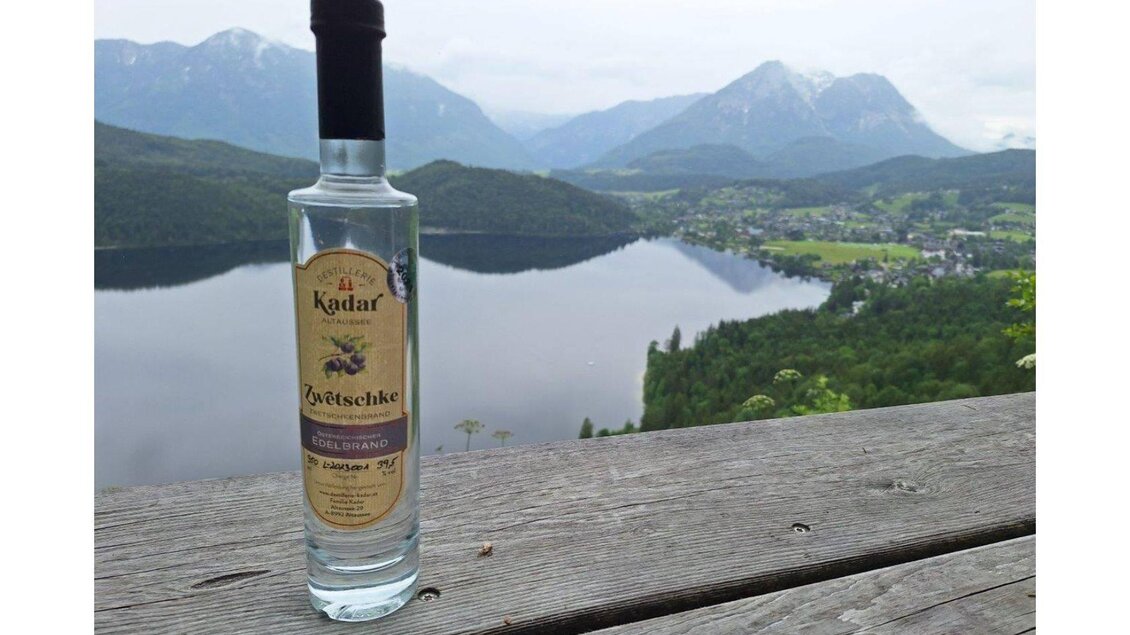 Eine Flasche Schnaps steht auf einem Holztisch mit Blick auf einen schönen See und die umliegenden Berge. Der Himmel ist bewölkt und die Landschaft ist grün und hügelig. | © Ernst Kadar
