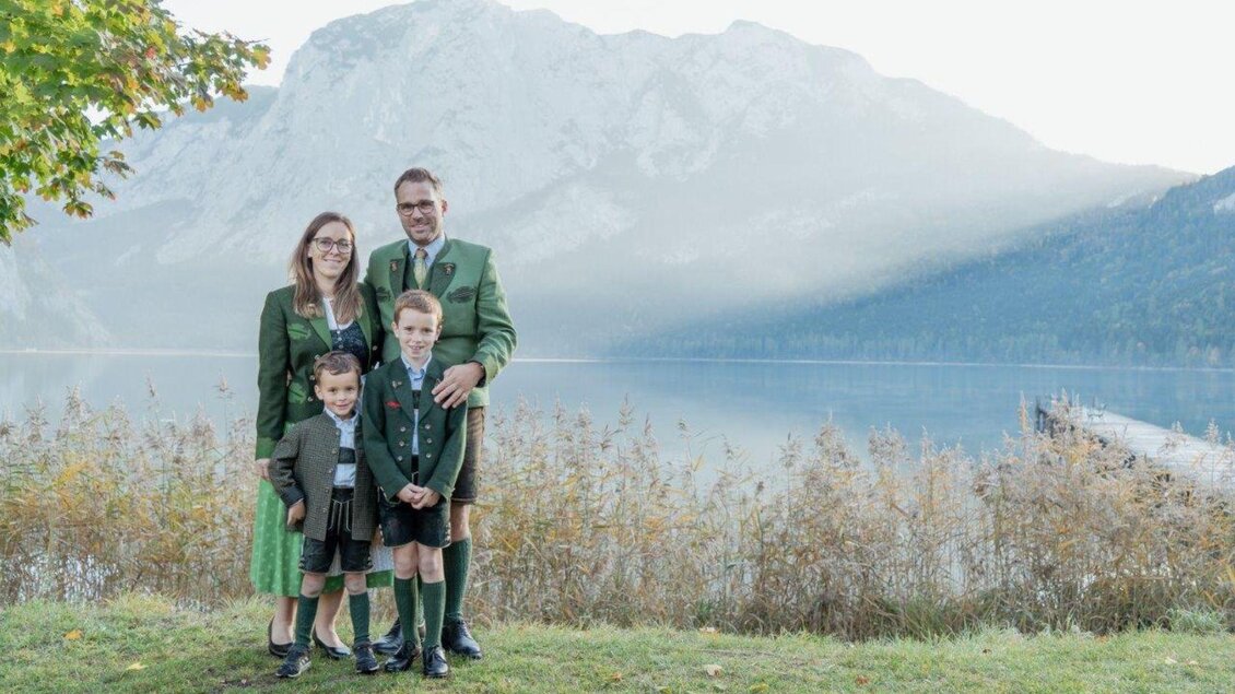 Eine Familie in traditionellen Trachten steht vor einem malerischen Bergsee. Im Hintergrund sind Wälder und Berge bei nebligem Wetter zu sehen. | © Christine Strutz - Lebensbilder
