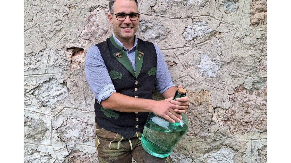 Ein Mann in traditioneller Tracht hält eine große grüne Flasche. Er steht vor einer rustikalen Steinwand. | © Ernst Kadar