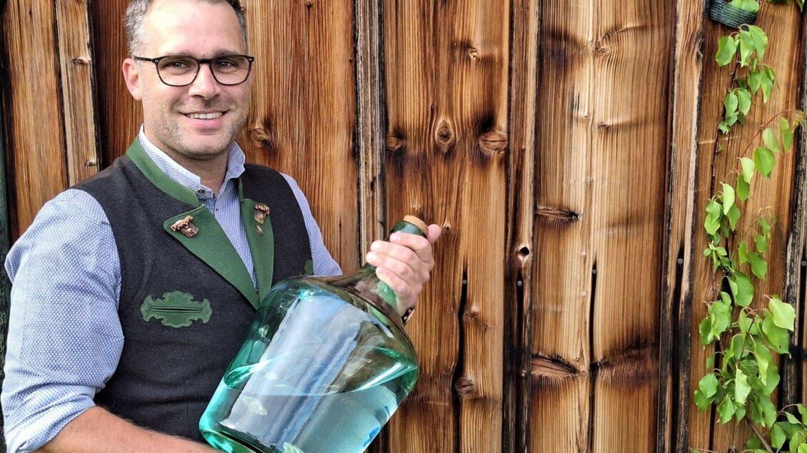 Ein lächelnder Mann in traditioneller Tracht hält eine große Glasflasche. Im Hintergrund sieht man eine Holzverkleidung und einige Pflanzen. | © Ernst Kadar