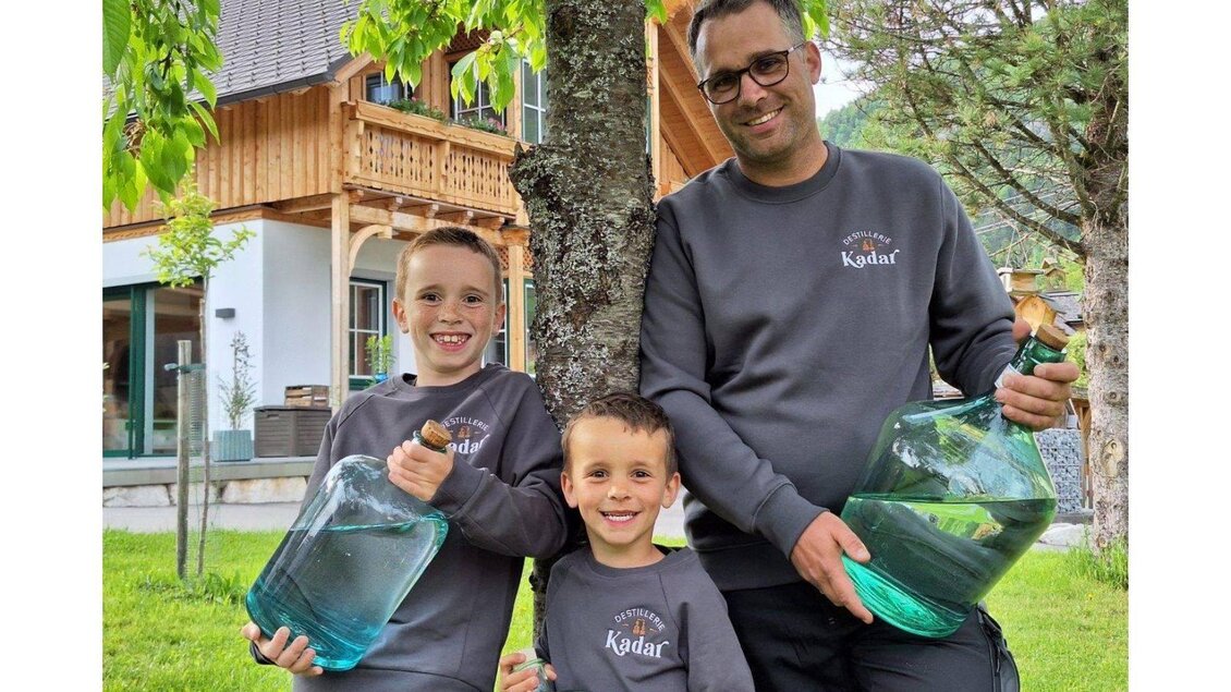 Ein Mann steht mit zwei Kindern unter einem Baum und hält große Glasflaschen. Alle tragen graue Sweatshirts und lächeln fröhlich. | © Ernst Kadar