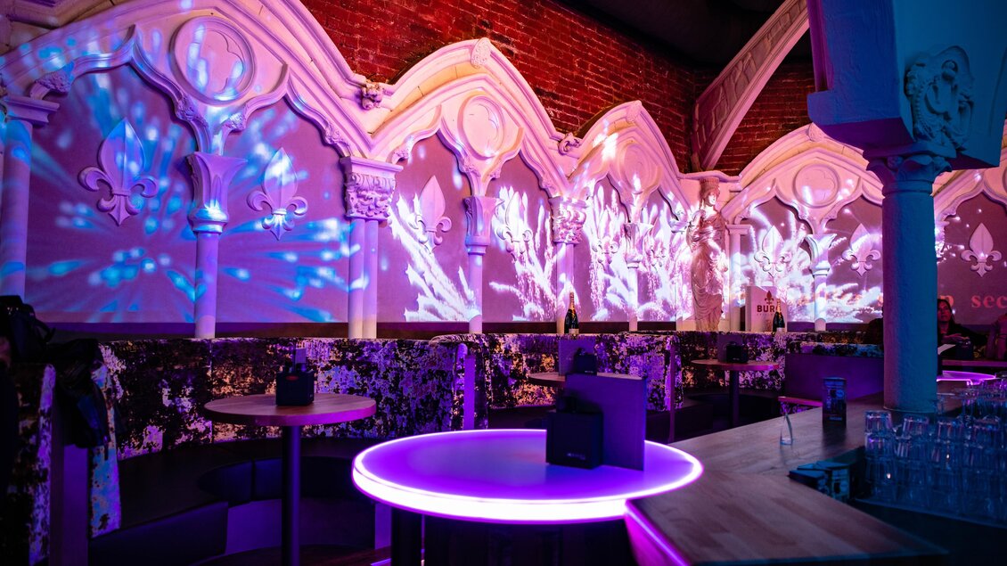 Eine stylische Bar mit bunten Lichtprojektionen an den Wänden. Der runde Tisch in der Mitte leuchtet in lila und schafft eine entspannte Atmosphäre. | © Die Burg