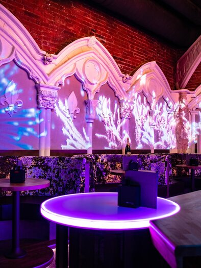 Eine stylische Bar mit bunten Lichtprojektionen an den Wänden. Der runde Tisch in der Mitte leuchtet in lila und schafft eine entspannte Atmosphäre. | © Die Burg