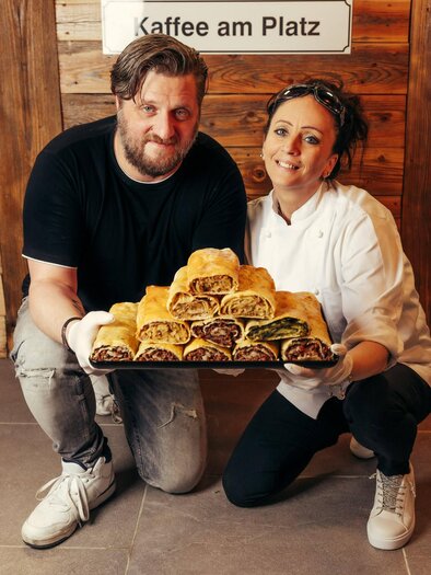 A cooking couple is holding a tray with various strudels. In the background, there is a sign with the inscription "Coffee at the table." | © Wolfgang Spekner