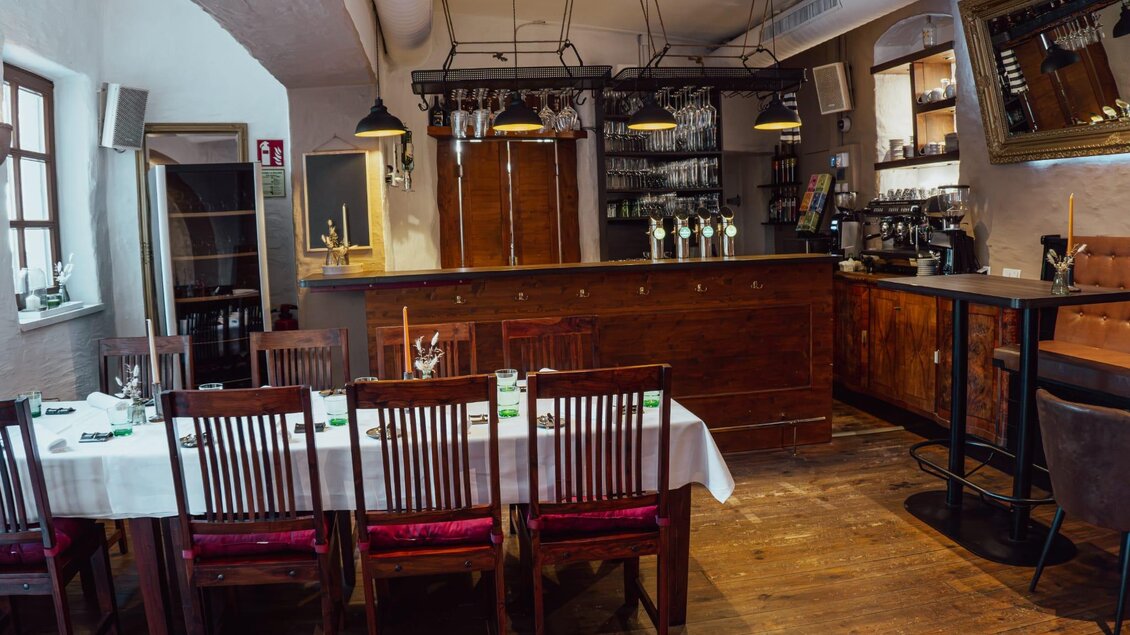 Ein gemütliches Restaurant mit Holzdekor und einer langen Tafelsituation. Eine Bar mit Gläsern und einem warmen Ambiente ist ebenfalls sichtbar. | © Oststeiermark Tourismus