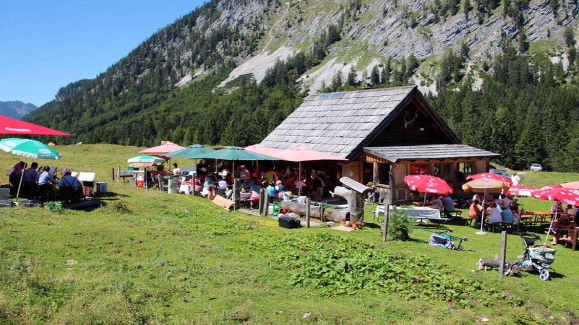 Eine gemütliche Berghütte umgeben von Wiesen und Bergen. Viele Menschen sitzen unter bunten Sonnenschirmen und genießen die Natur. | © Hotel DER HECHL