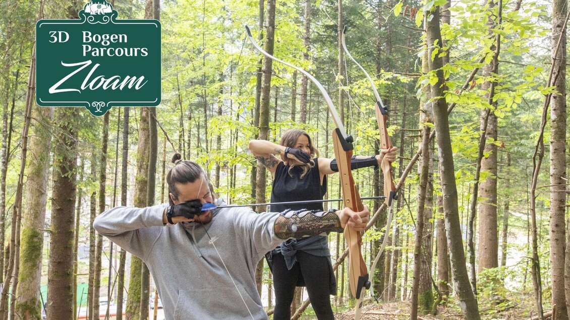 Zwei Personen schießen in einem Wald mit Bögen auf Ziele. Im Hintergrund sind hohe Bäume und ein Schild mit der Aufschrift "3D Bogen Parcours Zloam".