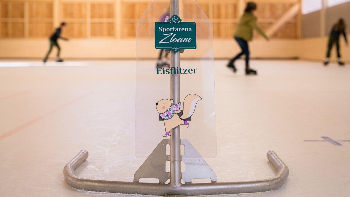 Ein Eislaufhilfe mit einem niedlichen Eichhörnchen-Motiv steht auf dem Eis. Im Hintergrund sind Eisläufer zu sehen, die fröhlich ihre Runden drehen. | © www.zloam.at