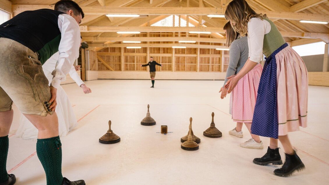 Eine Gruppe von Menschen spielt ein traditionelles Spiel in einer gepflegten Halle. Sie werfen Holzklötze auf eine glatte Fläche, während ein Spieler in der Mitte steht. | © www.zloam.at