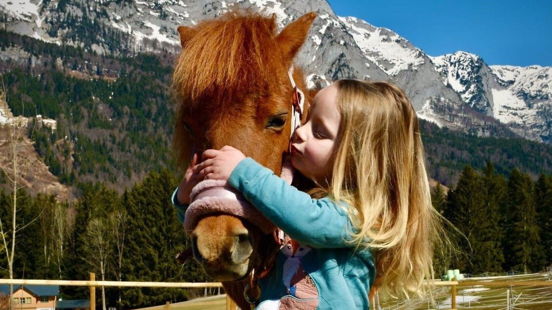 Ein Mädchen küsst ein Pony vor einer malerischen Berglandschaft. Im Hintergrund sind schneebedeckte Berge und grüne Wälder zu sehen. | © www.zloam.at