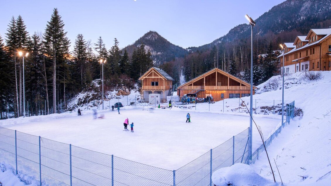 Eine wunderschöne Eislauffläche inmitten von schneebedeckten Bergen. Umgeben von Holzgebäuden und Tannenbäumen, genießen Menschen das Schlittschuhlaufen bei Dämmerung. | © www.zloam.at
