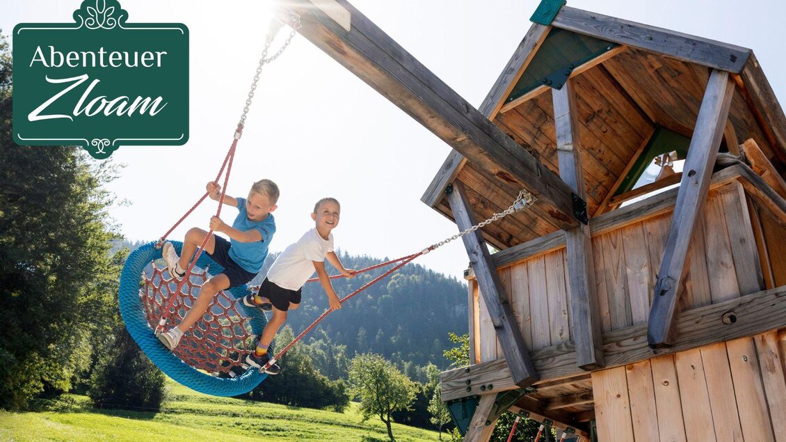 Zwei Kinder spielen auf einer schaukelnden Netzschaukel in einer grünen Umgebung. Im Hintergrund sieht man einen Spielturm und Bäume. | © www.zloam.at