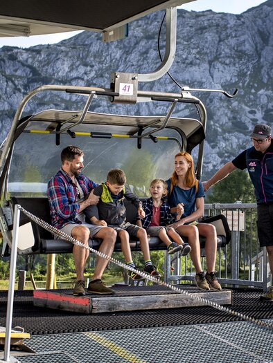 <p>Eine Familie sitzt am Sessellift mit der Berglandschaft der Tauplitzalm im Hintergrund. Sie bereiten sich auf den Ausstieg vor.</p> | © Die Tauplitz/Tom Lamm