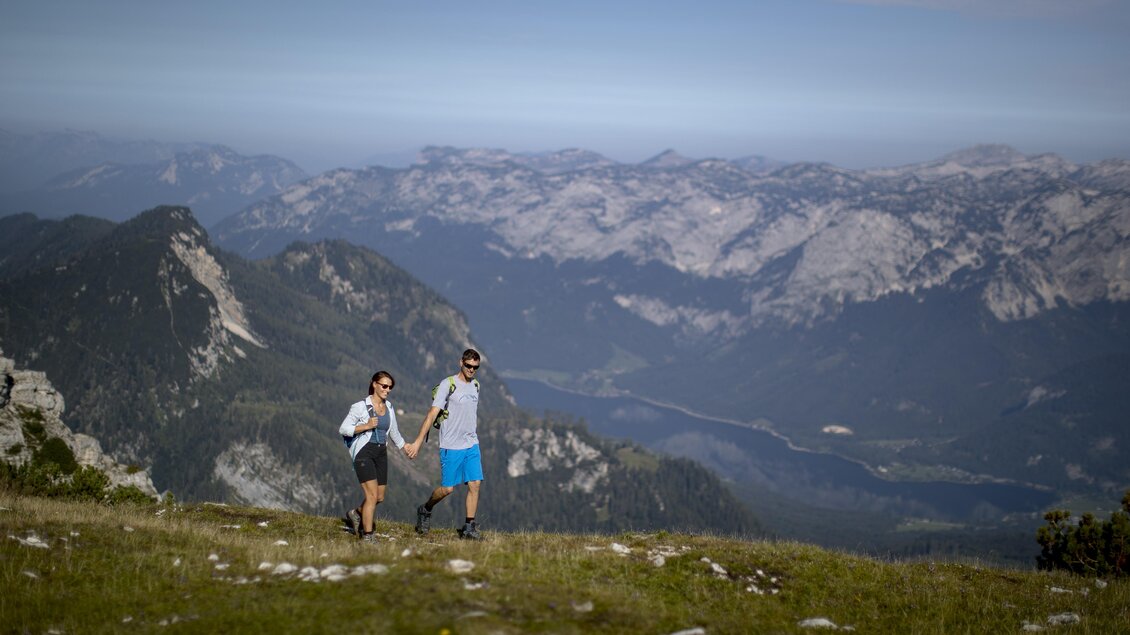 <p>Ein Paar wandert Hand in Hand auf dem Lawinenstein. Im Hintergrund ist diie beeindruckende Berglandschaft des Ausseerlandes mit dem Grundlsee und einem blauen Himmel zu sehen.</p> | © Die Tauplitz/Tom Lamm