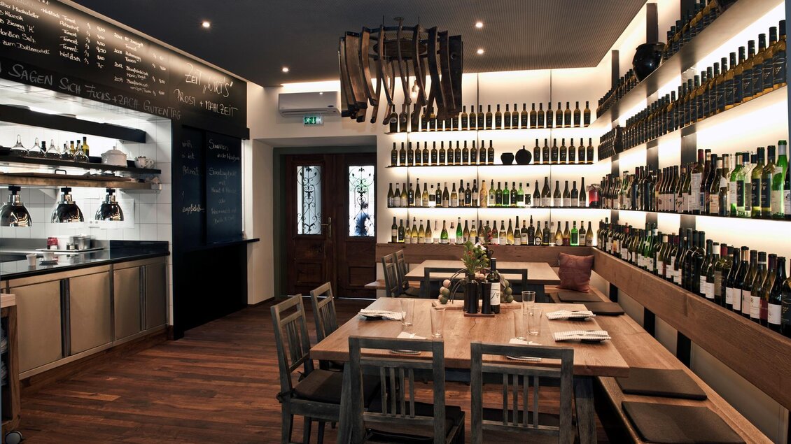 Ein modernes Restaurant mit Holztheken und vielen Weinflaschen an den Wänden. Große Tische und gemütliche Stühle laden zum Essen ein. | © Breitenthaler