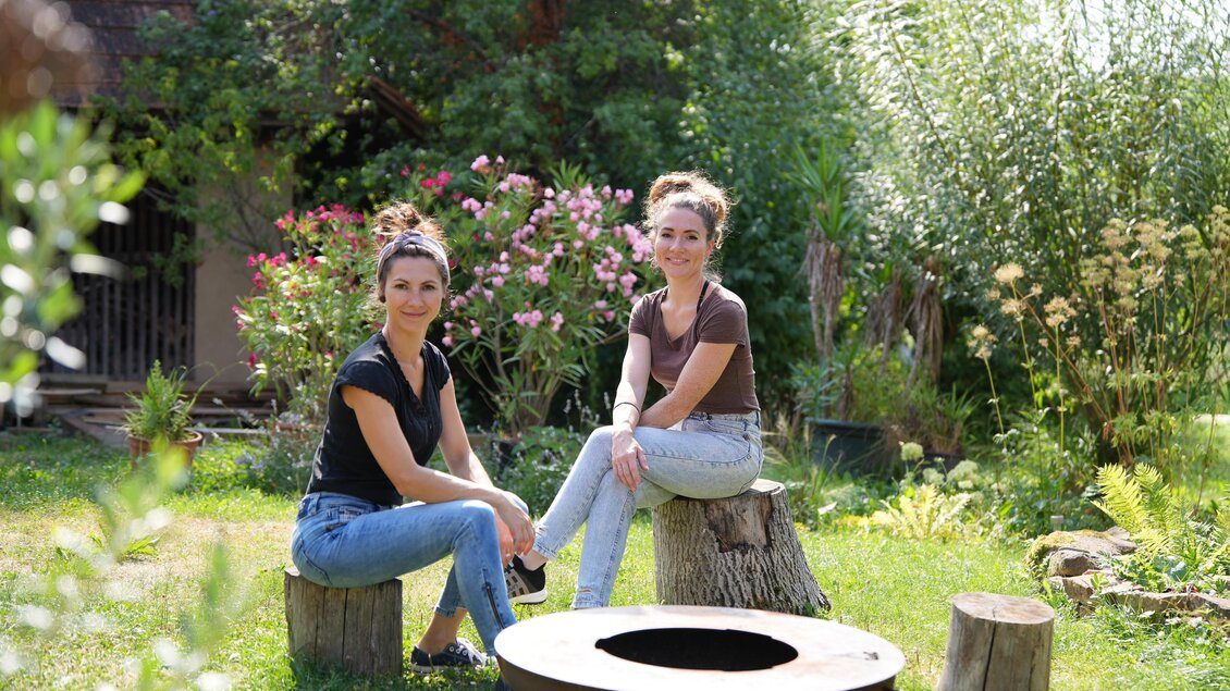 Zwei Frauen sitzen auf Baumstümpfen in einem Garten. Im Hintergrund sind Blumen und Bäume zu sehen.