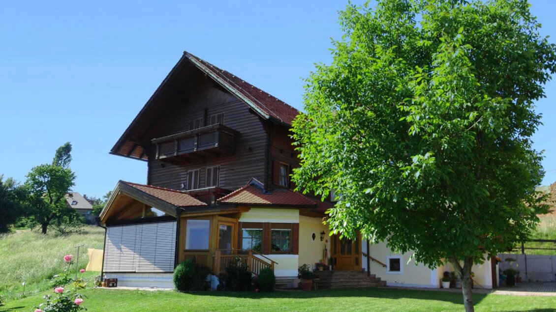 Ein charmantes Holzhaus umgeben von einem grünen Rasen und einem großen Baum. Der Himmel ist klar und blau. | © Gästehaus Klampfer