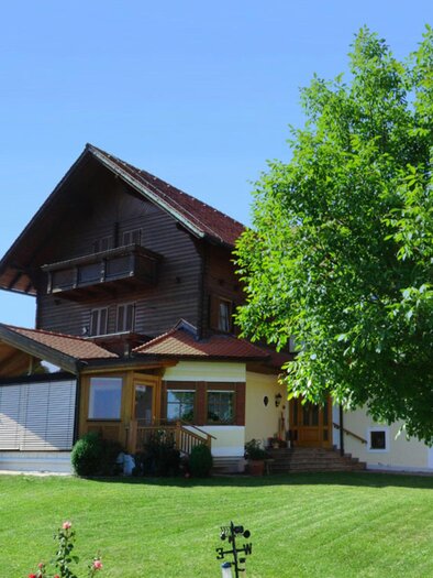 Ein charmantes Holzhaus umgeben von einem grünen Rasen und einem großen Baum. Der Himmel ist klar und blau. | © Gästehaus Klampfer