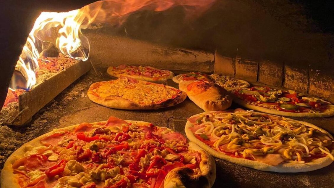 Mehrere belegte Pizzen backen neben offenem Feuer in einem Steinofen. | © pizzerija-pod-gradon