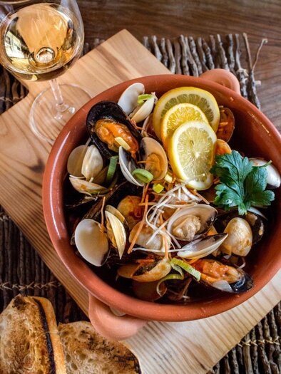 Eine Schüssel mit frischen Meeresfrüchten, garniert mit Zitronenscheiben und Kräutern. Neben dem Gericht steht ein Glas Weißwein und ein Stück Brot auf einem Holzbrett. | © Don Camillo