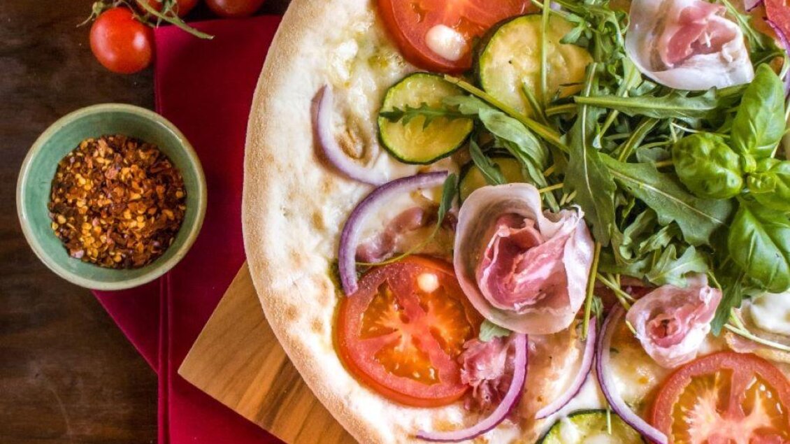 Eine frisch belegte Pizza mit Tomaten, Gurken und Rucola. Daneben befindet sich eine Schüssel mit roten Pfefferflocken und einige Cherrytomaten. | © Don Camillo