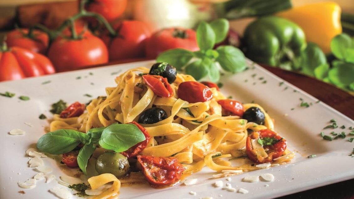 Ein Teller Pasta mit frischen Tomaten, Oliven und Basilikum. Im Hintergrund sind verschiedene Gemüsesorten zu sehen. | © Don Camillo