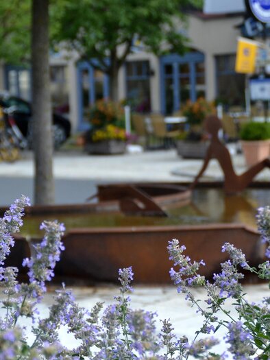 <p>A picturesque garden scene with purple flowers in the foreground and large, rustic water containers in the background. In the background, trees and a building with windows can be seen. Village well in Bad Blumau.</p> | © Kurkommission Bad Blumau