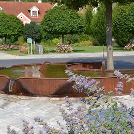 Eine moderne Brunnenanlage umgeben von Bäumen und Blumen. Das Wasser fließt sanft in eine runde, metallene Schale. | © Kurkommission Bad Blumau