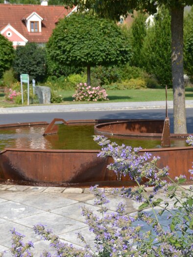 Eine moderne Brunnenanlage umgeben von Bäumen und Blumen. Das Wasser fließt sanft in eine runde, metallene Schale. | © Kurkommission Bad Blumau