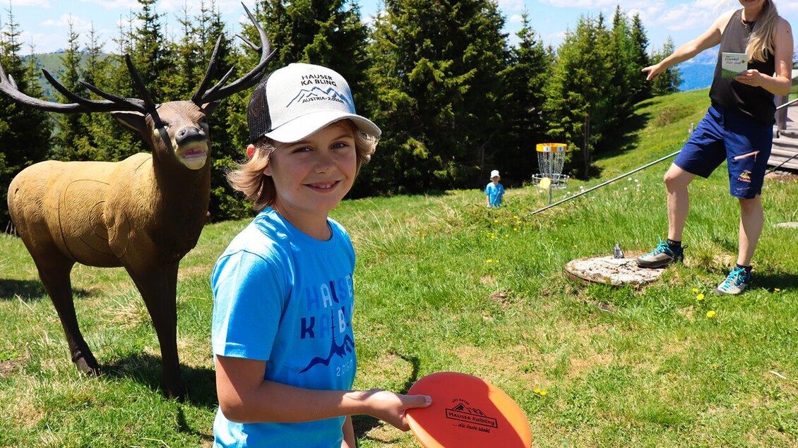Ein Junge mit einem Frisbee steht lächelnd in der Natur. Im Hintergrund sieht man eine Frau, die auf eine Zielscheibe zeigt, sowie ein Tiermodell. | © Hauser Kaibling