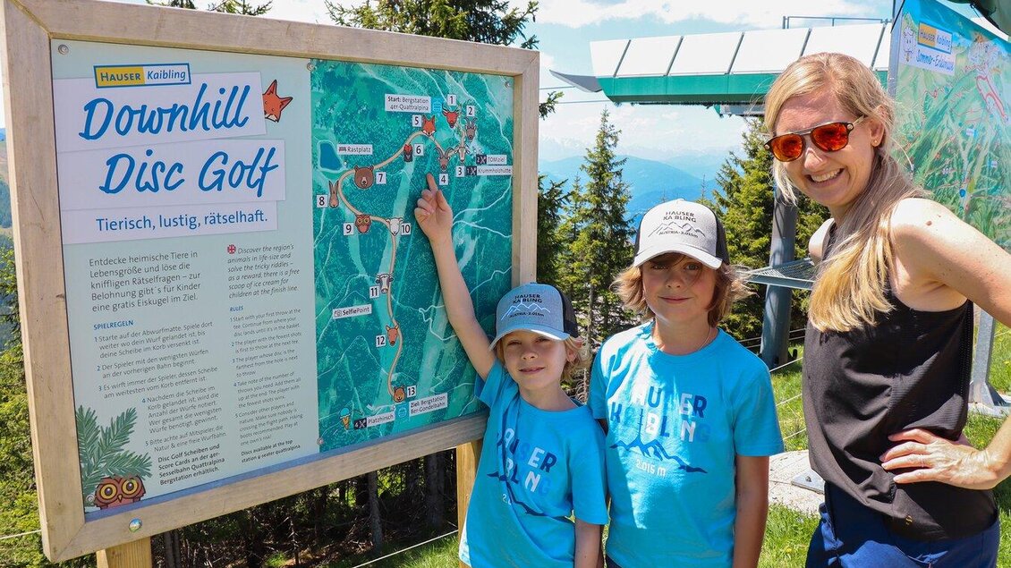 Eine Mutter steht mit zwei Jungen vor einem Informationsschild über Disc Golf. Sie zeigen auf die Karte und genießen einen sonnigen Tag in der Natur. | © Hauser Kaibling